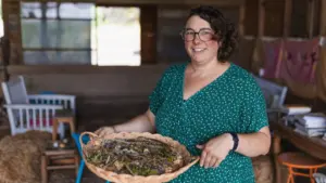 Une femme souriante aux cheveux courts tenant un plateau en osier rempli de plantes séchées.