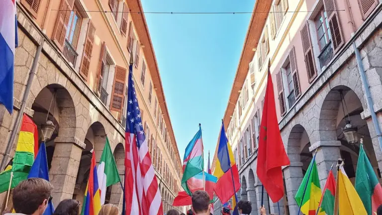 Une foule portant divers drapeaux nationaux marche dans une rue bordée de bâtiments historiques.