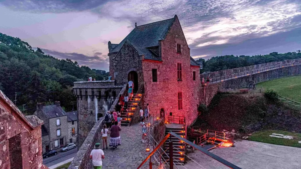 Un groupe de visiteurs monte les escaliers en pierre d'un château médiéval illuminé au crépuscule.