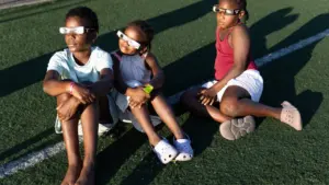 Trois enfants assis sur une pelouse synthétique portant des lunettes de protection pour éclipse solaire.