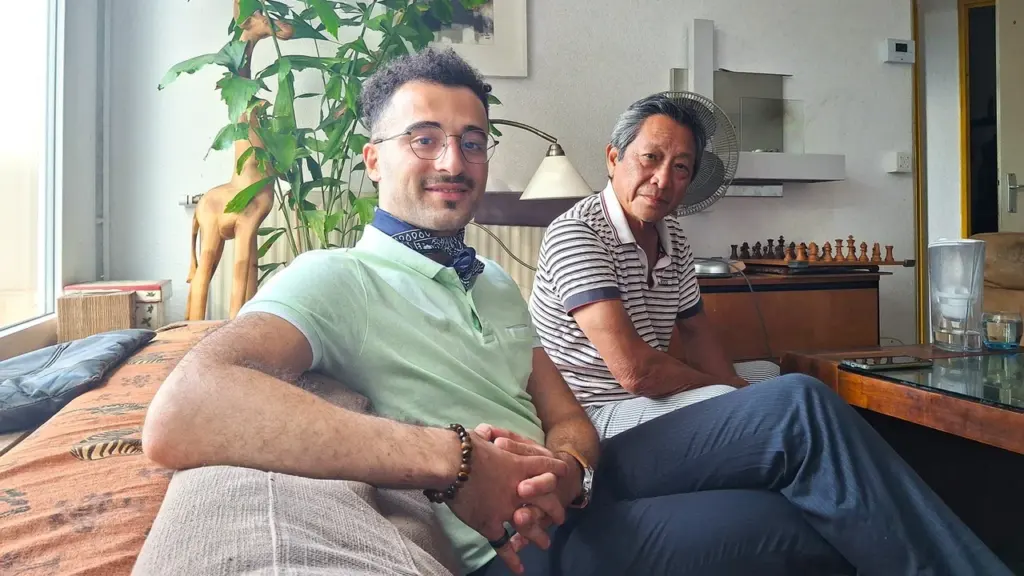 Deux hommes assis côte à côte sur un canapé dans un salon lumineux et chaleureux.