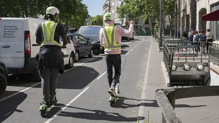 Deux personnes en trottinettes électriques circulent sur une piste cyclable dans une rue parisienne.