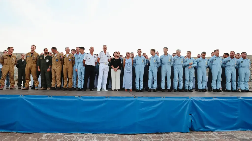 Un groupe de pilotes militaires et deux femmes posent ensemble sur une estrade bleue extérieure.