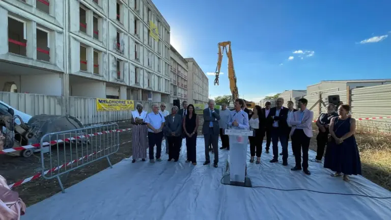 Un groupe de personnes devant un grand bâtiment en démolition avec une pelleteuse jaune.