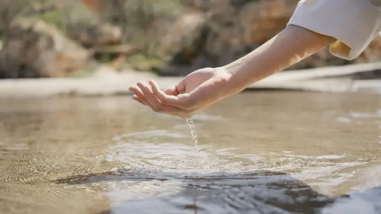Une main ouverte effleurant la surface d'une eau claire et calme en extérieur.